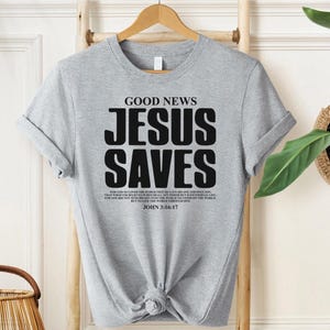 May include: Gray t-shirt with the text "GOOD NEWS JESUS SAVES" in black. The shirt also includes a Bible verse from John 3:16-17.