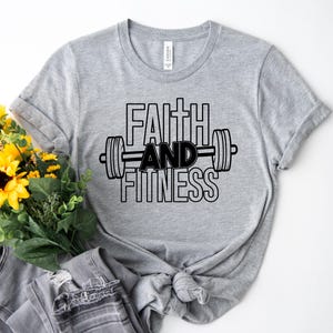 Może przedstawiać: Szara koszulka z czarnym napisem "Faith and Fitness" i grafiką sztangi.