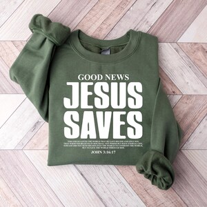 May include: Green sweatshirt with the text "GOOD NEWS JESUS SAVES" in white. The text is in a bold font. Below the text is a Bible verse: "For God so loved the world that he gave his one and only Son, that whoever believes in him shall not perish but have eternal life. For God did not send his Son into the world to condemn the world, but to save the world through him. JOHN 3:16-17"