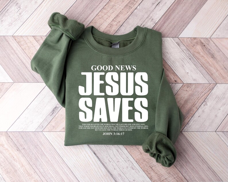 May include: Green sweatshirt with the text "GOOD NEWS JESUS SAVES" in white. The text is in a bold font. Below the text is a Bible verse: "For God so loved the world that he gave his one and only Son, that whoever believes in him shall not perish but have eternal life. For God did not send his Son into the world to condemn the world, but to save the world through him. JOHN 3:16-17"