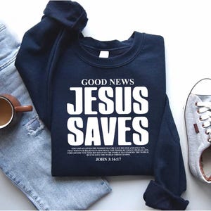 May include: A navy blue sweatshirt with the text "GOOD NEWS JESUS SAVES" in white. The text is in a bold, blocky font. Below the text is a Bible verse from John 3:16-17.