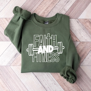 Może przedstawiać: Zielona bluza z napisem "Faith and Fitness" w białym kolorze i grafiką białej sztangi.