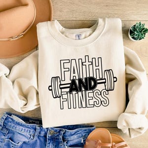 Może przedstawiać: Kremowa bluza z nadrukiem "Faith and Fitness" w kolorze czarnym, z graficznym przedstawieniem sztangi.
