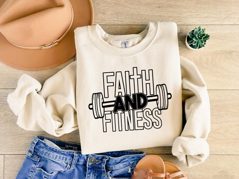 Może przedstawiać: Kremowa bluza z nadrukiem "Faith and Fitness" w kolorze czarnym, z graficznym przedstawieniem sztangi.