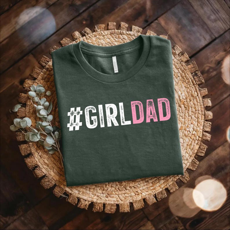 May include: A dark green t-shirt with the text "#GIRLDAD" in white and pink letters. The letters have a distressed, vintage look.