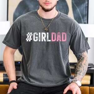 May include: A dark gray t-shirt with the text "#GIRLDAD" printed in white and pink letters.