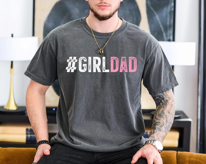 May include: A dark gray t-shirt with the text "#GIRLDAD" printed in white and pink letters.