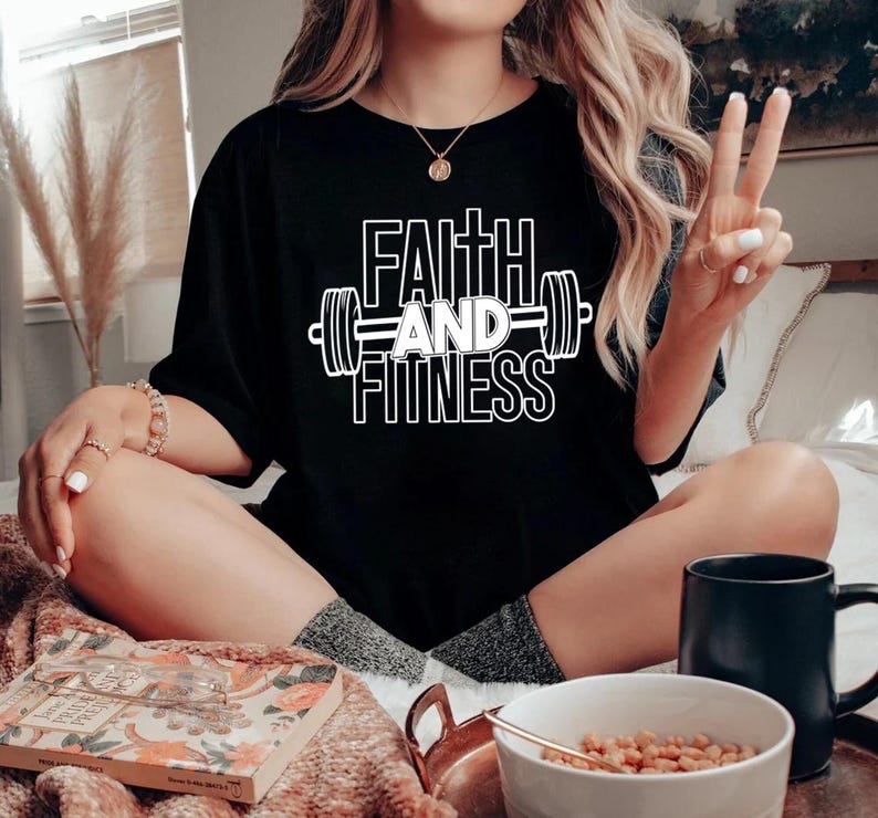Może przedstawiać: Czarny t-shirt z napisem "Faith and Fitness" w białym kolorze, z grafiką sztangi. Koszulkę nosi osoba siedząca na kanapie.