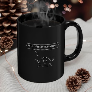 Op de afbeelding: Zwarte koffiemok met een wit ontwerp van een krab met de tekst "< Hello fellow Rustaceans! >" op een zwarte achtergrond.