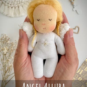 May include: A handmade doll with a light skin tone, blonde hair, and a white outfit is held in cupped hands. The doll has closed eyes and rosy cheeks. The text "ANGEL ALLURA" and "Doll Pattern and Instructions" are displayed.