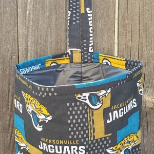 May include: A black fabric tote bag with a Jacksonville Jaguars design. The bag features a repeating pattern of the team's logo, which includes a jaguar head with teal, gold, and black colors. The bag has a black fabric handle.