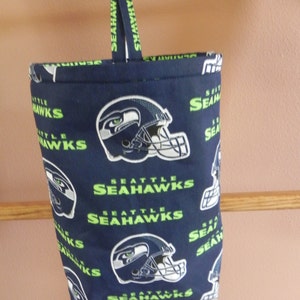 May include: A navy blue fabric bag featuring the Seattle Seahawks logo and text in green and white. The bag has a loop handle with the team name printed on it. The bag is hanging from a wooden bar.