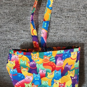 May include: A colorful tote bag featuring a repeating pattern of cartoon cats in various colors. The bag has a rectangular shape with a wide opening and two matching handles. The handles and top trim have a vibrant, abstract design.