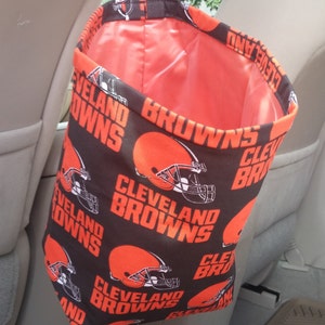 May include: A black and orange fabric car trash can with a hanging strap. The fabric has a repeating pattern of the Cleveland Browns logo, which includes a football helmet and the words "Cleveland Browns".
