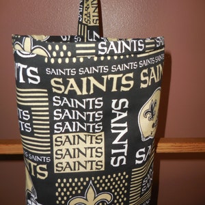 May include: A black and gold fabric bag with a Saints logo and the word "Saints" repeated multiple times. The bag has a black strap with the word "Saints" printed on it.