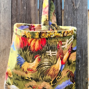 May include: A green fabric tote bag with a yellow handle. The bag features a colorful print of roosters, pumpkins, and sunflowers.