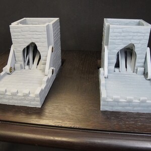 May include: Two gray 3D printed dice towers with a stone-like texture. Each tower has a hinged opening with a wooden drawbridge design. The towers are on a dark wood surface.