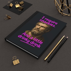 May include: A black hardback journal featuring a portrait of a bearded man in a classical style. The cover displays the bright pink text "I require snackies. And a little drinky drink" printed above and below the central figure's head.