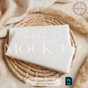 May include: A white chocolate bar mockup on a woven straw placemat with a beige background. The text "Chocolate Bar Mock Up" is written in white on the bar.