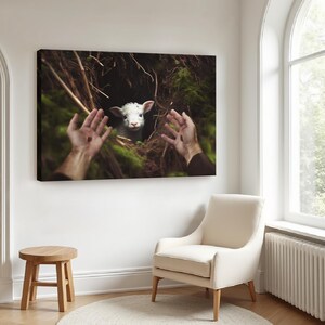 May include: A canvas print depicting a lamb in a dark, mossy setting, with outstretched hands framing the scene. The artwork hangs on a white wall, above a white chair and a wooden stool, creating a serene atmosphere.