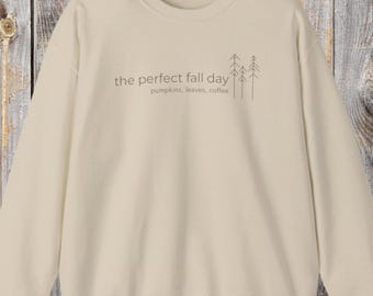 Puede incluir: Sudadera de color crema con el texto "the perfect fall day" y las palabras "pumpkins, leaves, coffee" debajo. Un gráfico minimalista de árbol está a la derecha del texto. La sudadera está hecha de un material suave.