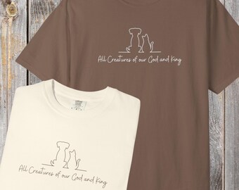 Könnte beinhalten: Zwei T-Shirts, eines cremefarben und eines braun, werden gezeigt. Jedes hat eine Strichzeichnung eines Hundes und einer Katze mit dem Text "All Creatures of our God and King" darunter. Die T-Shirts sind aus weichem Material.