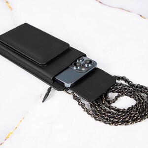 May include: A black leather phone case with a chain strap. The case has a pocket on the front and a slot for a phone on the back. The phone is not visible in the image.