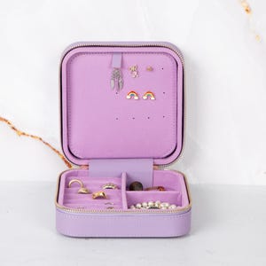 Puede incluir: Estuche de viaje para joyas lila abierto con cierre de cremallera. El interior presenta un forro suave de color lila claro con compartimentos para anillos, pendientes y collares. El estuche contiene varios pendientes, anillos y un collar de perlas.