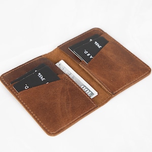 May include: A brown leather card holder wallet featuring two interior pockets and a central compartment. The wallet contains two black credit cards with white text and a one hundred dollar bill tucked into the centre slot, showing visible stitching.