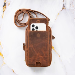 May include: A brown leather crossbody bag with a smartphone in the front pocket. The bag has a long, adjustable strap and a small zipper pocket on the side. The leather has a distressed finish, and the bag is set against a marble background.