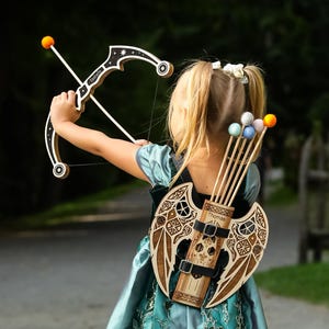 May include: A wooden toy bow and arrow set with a quiver on the back. The bow is white and brown with a star design. The quiver is brown with a decorative design and holds several arrows with colorful tips.