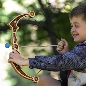 Puede incluir: Un juego de arco y flecha de juguete de madera. El arco es de color marrón claro con adornos de estrellas y círculos. Una bola azul está unida a la cuerda del arco. El niño sostiene el arco y la flecha, apuntando a un objetivo.