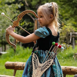 May include: A young girl aims a wooden bow and arrow. The bow has decorative carvings. She wears a teal dress and has a quiver with colorful arrows. The scene takes place outdoors on a wooden bridge.