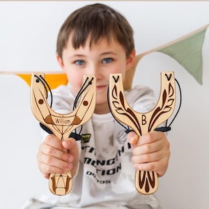 May include: Two wooden slingshots with black bands are held up. One has the name "William" engraved, the other has the letter "B". The text at the top reads "PERFECT GIFT FOR LITTLE ONES ADVENTURERS". The slingshots have a light brown color with decorative leaf patterns.