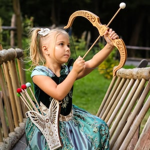 May include: A young girl in a teal and green dress holds a wooden bow and arrow. The bow has decorative carvings. A quiver with colorful arrows is attached to her side. The scene is set outdoors with a wooden fence in the background.