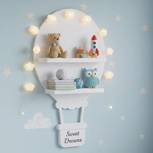 May include: A white hot air balloon-shaped shelf with two levels, adorned with string lights, is mounted on a light blue wall. The shelf holds a teddy bear, a rocket, books, and an owl figurine. The words "Sweet Dreams" are on the basket.