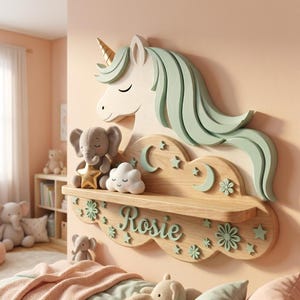 May include: A wooden unicorn-shaped shelf with a gold horn and mint green mane. The shelf is decorated with stars, moons, and flowers, and has the name "Rosie" in mint green script. A plush elephant, star, and cloud sit on the shelf.