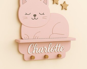 May include: A pink wooden cat-shaped shelf with the name "Charlotte" in white script. The shelf has three wooden pegs below and two wooden star decorations above. The cat has closed eyes.