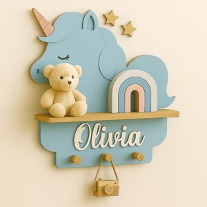 May include: A light blue wooden shelf with a unicorn design, a teddy bear, and a rainbow. The shelf has the name "Olivia" in white script, three pegs, and a small wooden camera hanging below. Two gold stars are above the unicorn.