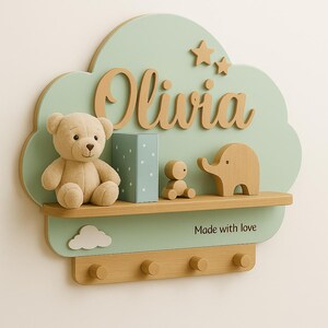 Puede incluir: Estantería de pared en forma de nube verde menta con el nombre "Olivia" en letras de madera. Cuenta con un osito de peluche, una pequeña caja azul y figuras de madera. La estantería incluye ganchos y la frase "Made with love".