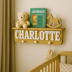 Könnte beinhalten: Ein Holzregal mit dem Namen "CHARLOTTE" in weißen Buchstaben. Das Regal enthält einen Teddybären, ein Buch mit dem Titel "TIERE" und ein Schaukelpferd aus Holz. Darunter befinden sich drei Haken und ein kleiner beiger Beutel.