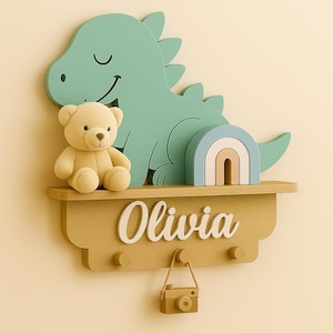 Puede incluir: Estantería de pared de madera con un dinosaurio verde sonriente, un oso de peluche y un arcoíris. La estantería incluye el nombre "Olivia" y una pequeña cámara de madera colgando debajo. Incluye tres pequeños ganchos.