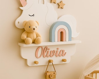 May include: A white wooden unicorn-themed shelf with a shelf, hooks, and a name cutout. A teddy bear, a rainbow, and a wooden camera are on the shelf. The name "Olivia" is displayed in pink script.