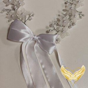 Könnte beinhalten: Eine weiße Satin Schleife mit vier langen Bändern, gebunden an einem weißen Blumen-Haarreif mit Perlen-Akzenten.