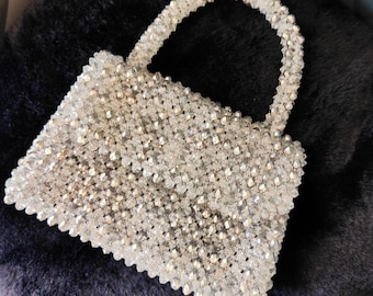 May include: A small, rectangular handbag covered in clear, sparkling beads. The handle is also made of the same beads. The bag is sitting on a dark, textured surface, creating a contrast that highlights the bag's shimmering appearance. The bag is a fashion accessory.