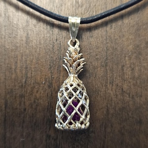 May include: A silver-colored pineapple pendant with a dark purple stone center, suspended from a black cord. The pendant has a detailed, openwork design, and the pineapple top is textured. The background is a wooden surface.