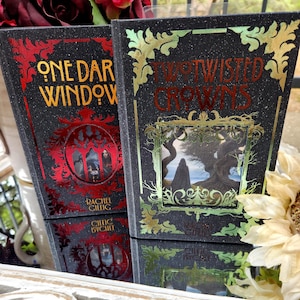 May include: Two hardcover books with black covers and gold and red accents. The book on the left is titled "One Dark Window" and the book on the right is titled "Twisted Crowns". Both books have decorative frames and the author's name.