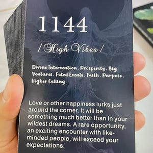 Puede incluir: Una baraja de cartas de oráculo con un fondo azul oscuro. La carta en foco muestra el número "1144" y las palabras "High Vibes". El texto en la tarjeta incluye "Divine Intervention, Prosperity, Big Ventures, Fated Events, Faith, Purpose, Higher Calling."