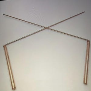 May include: A pair of copper-colored metal dowsing rods, with two straight handles and two crossed rods. The rods are designed to intersect at a point, creating an X shape. The handles are cylindrical.