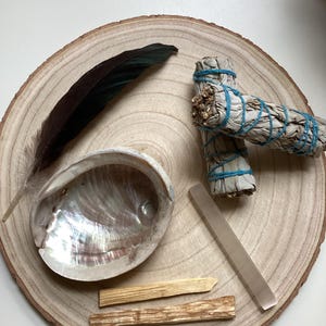 May include: A wooden tray with a white sage smudge stick, a black feather, a seashell, two wooden sticks, and a clear quartz crystal.
